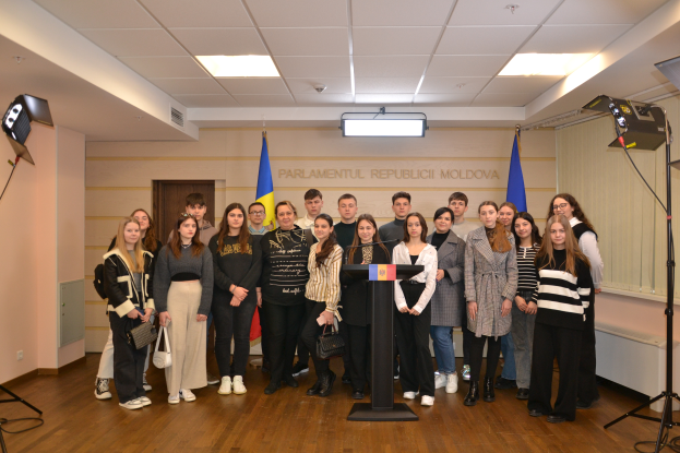 Eine Gruppe von Menschen steht vor einem Podium mit einem Mikrofon, umgeben von Fahnen, einer Wand mit dem Text "Moldova Jugendkongress 2018", einer Tür, einem Dach mit Deckenleuchten und Ständern mit Leuchten.
