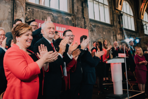 Eine Gruppe von Menschen, die vor einem Publikum applaudieren, mit einem Podium, einem Mikrofon und einer Texttafel auf der rechten Seite und Stühlen, einer Fahne, einer Wand, Fenstern und Lichtern im Hintergrund.