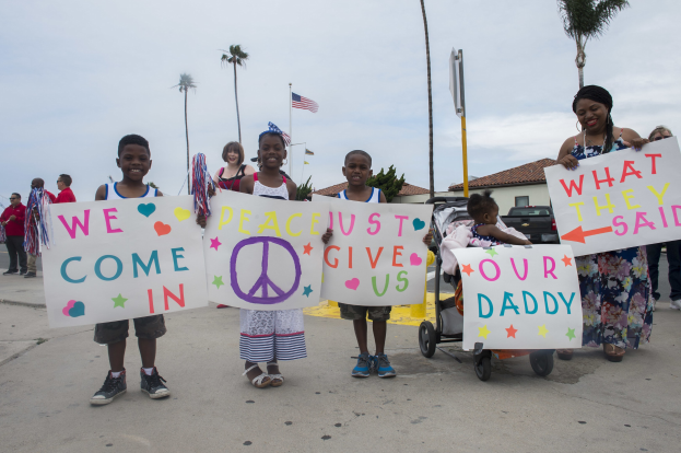 Eine Gruppe von Kindern hält Schilder und steht vor einem Gebäude, während eine Frau einen Kinderwagen mit einem Baby schiebt; auf einem Schild steht: "Wir kommen in Frieden, gebt uns unseren Daddy."