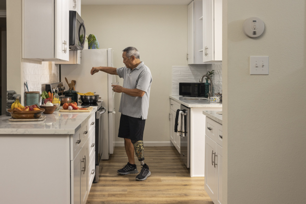 Ein Mann steht in einer Küche mit weißen Schränken und hält ein Glas in der Hand, während verschiedene Gegenstände wie Obst, Gemüse, Schüsseln und Löffel auf einer Arbeitsplatte zu seiner Linken angeordnet sind.