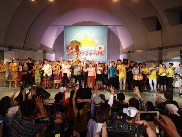 Gruppe von Menschen auf einer Bühne mit Kameras, Handys und Musikinstrumenten während einer Festival-Eröffnung mit Bannern, Wänden und Deckenlampen im Hintergrund.