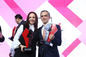 Zwei Personen auf einer Bühne, die lächeln und Pokale halten, mit einem Bildschirm im Hintergrund.