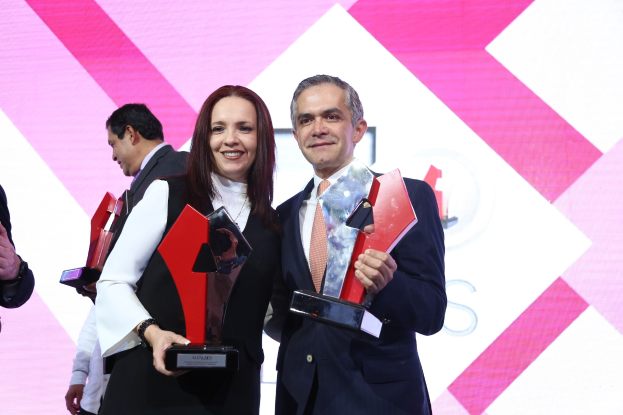 Zwei Personen auf einer Bühne, die lächeln und Pokale halten, mit einem Bildschirm im Hintergrund.