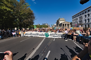 Eine große Gruppe von Menschen marschiert auf einer Straße in Berlin, hält Schilder und Plakate, während rechts eine Person mit einer Kamera und einem Mikrofon zu sehen ist, links Bäume und Gebäude und ein klarer Himmel.