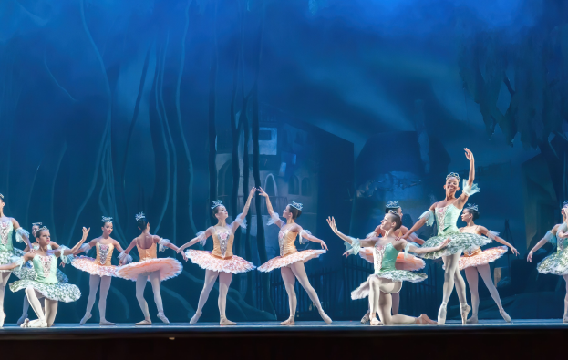 Eine Gruppe von Balletttänzerinnen in bunten Kostümen, die *Der Nussknacker* auf der Bühne des Royal Opera House tanzen, bewegen sich synchronisiert gegen einen blauen Hintergrund.