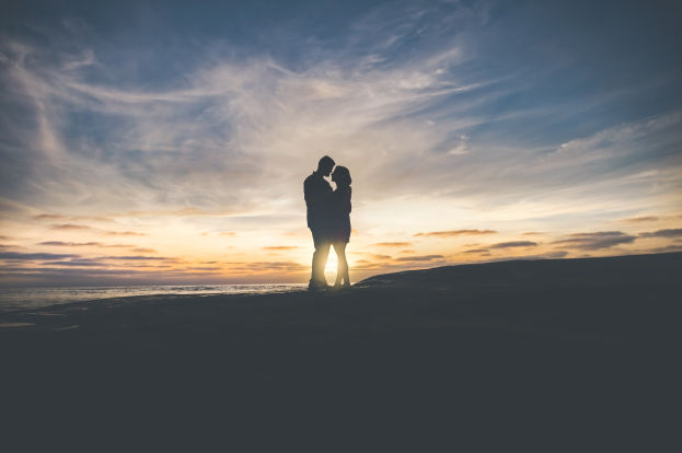 Silhouette eines Paares, das sich auf einem Strand bei Sonnenuntergang umarmt und küsst, mit leuchtendem Orange, Pink und Purpurhimmel und tiefem Blauem Wasser.