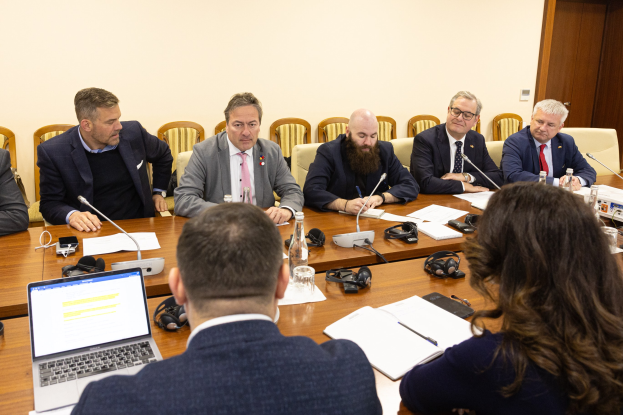 Eine Gruppe von Menschen, wahrscheinlich Mitglieder des Europäischen Parlaments, sitzt um einen Tisch mit Laptops, Büchern, Papieren, Flaschen, Gläsern, Headsets, Mikrofonen mit Ständern und Mobiltelefonen in einem Konferenzraum mit einer Wand und einer Tür im Hintergrund.