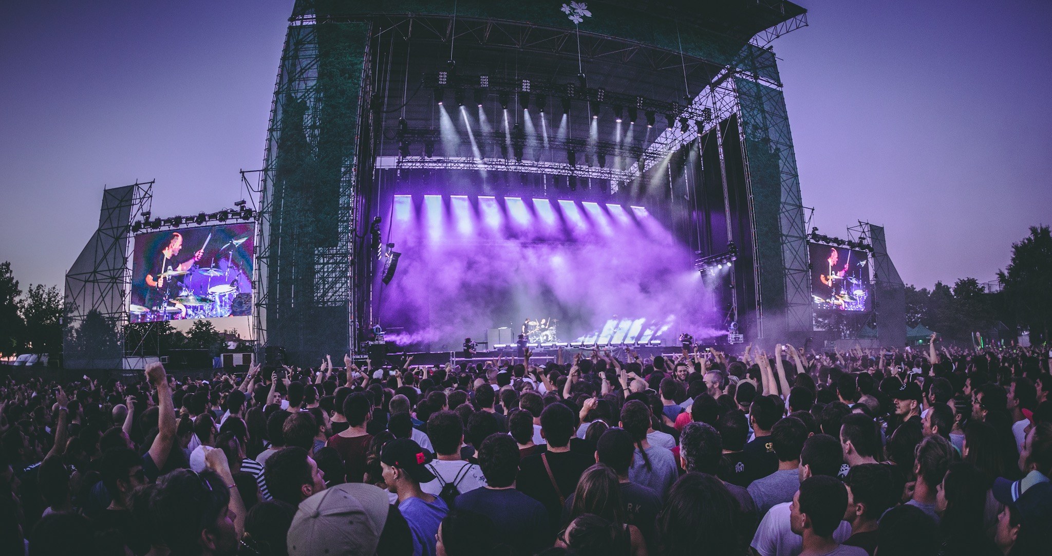 Eine große Menschenmenge bei einem Musikfestival schaut Künstlern auf einer hell erleuchteten Bühne mit Bildschirmen zu, die vor einem Gebäude, Bäumen und einem klaren blauen Himmel aufgebaut ist.