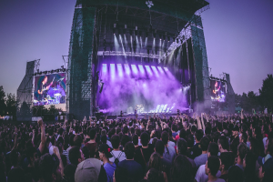 Eine große Menschenmenge bei einem Musikfestival schaut Künstlern auf einer hell erleuchteten Bühne mit Bildschirmen zu, die vor einem Gebäude, Bäumen und einem klaren blauen Himmel aufgebaut ist.