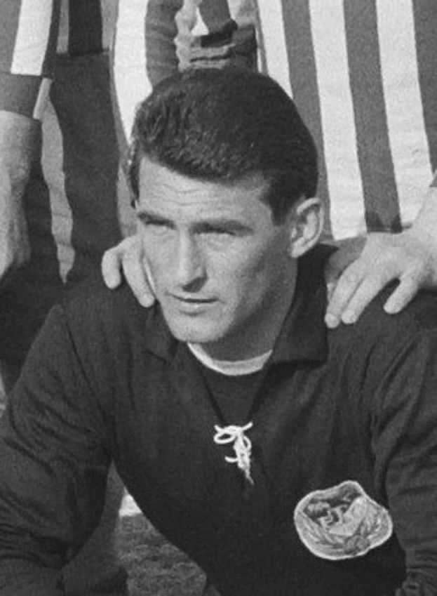 Schwarzes und weißes Foto eines Mannes, der auf einem Rasenfußballfeld sitzt und ein T-Shirt und eine Kette trägt, während zwei Personen hinter ihm stehen.