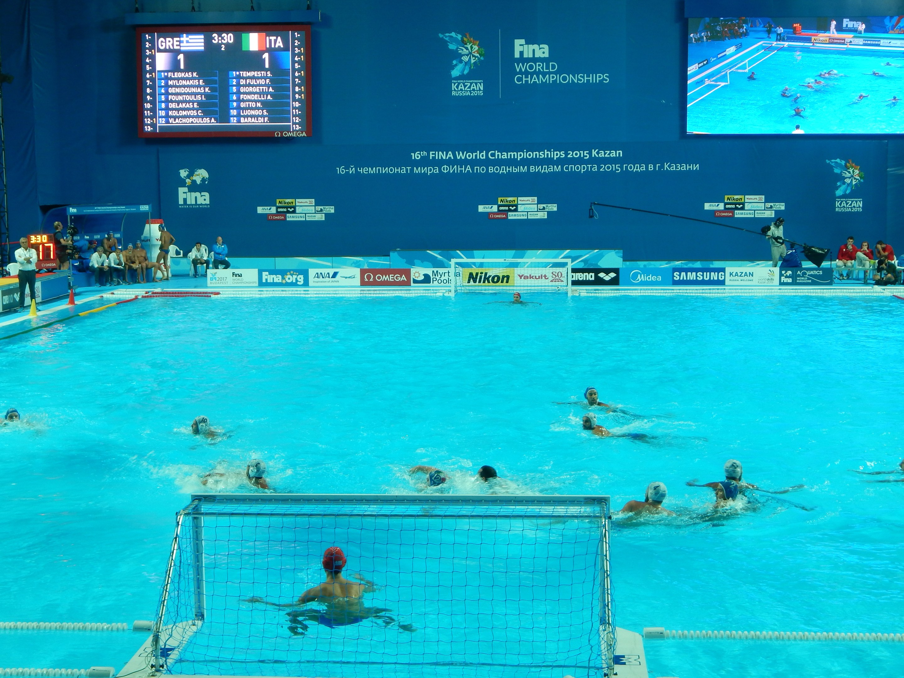 Gruppe von Athleten, die Wasserball in einem Pool während der FINA-Weltmeisterschaften 2016 in Kazan spielen, mit Zuschauern auf der linken Seite und einem Bildschirm im Hintergrund, der Werbung anzeigt.