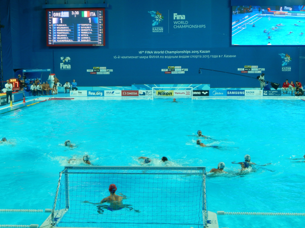 Gruppe von Athleten, die Wasserball in einem Pool während der FINA-Weltmeisterschaften 2016 in Kazan spielen, mit Zuschauern auf der linken Seite und einem Bildschirm im Hintergrund, der Werbung anzeigt.
