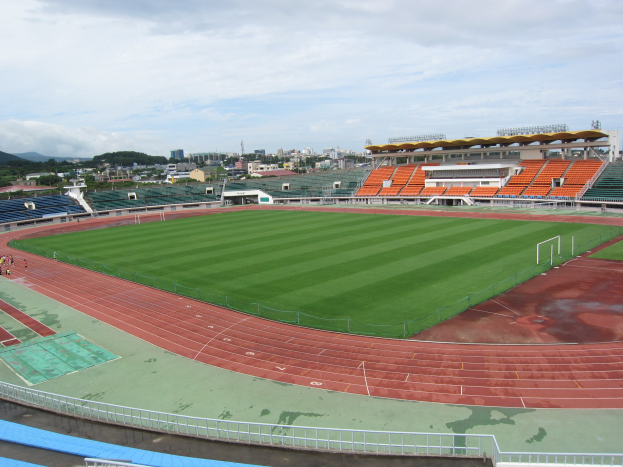 Großes Stadion mit einem Fußballfeld, umgeben von Gebäuden, Bäumen, Hügeln und einem klaren blauen Himmel, mit einigen Menschen und saftigem Grün.