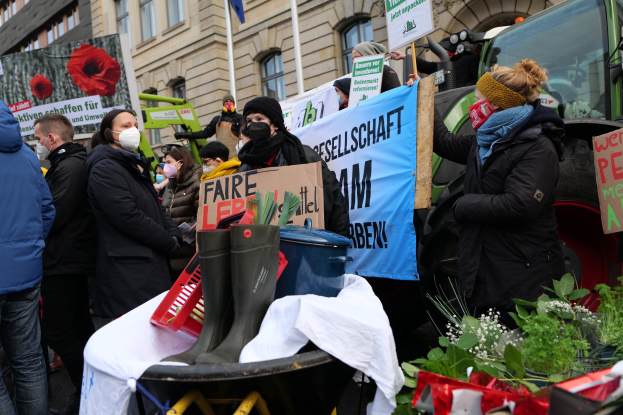 Eine Gruppe von Menschen mit Masken und Protestschildern vor einem Lastwagen, mit einem Tisch mit verschiedenen Gegenständen, Topfpflanzen und einem Gebäude mit Fahne im Hintergrund.