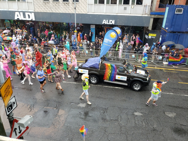 Eine Gruppe von Menschen in bunter Kleidung, die eine Straße in einer Parade entlanggehen, mit einem Auto in der Mitte, einige halten Schirme, und Gebäude mit Fenstern im Hintergrund.