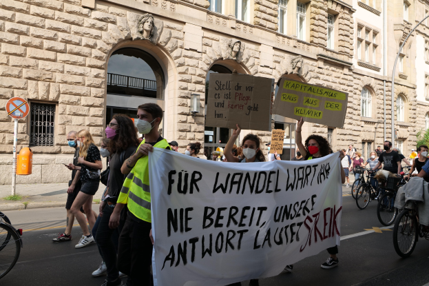 Eine Gruppe von Menschen, die auf einer Protestdemo durch die Stadt marschieren, einige halten Schilder und andere fahren Fahrräder, im Hintergrund ein historisches Gebäude mit Rundbogenfenstern, Säulen und Skulpturen.
