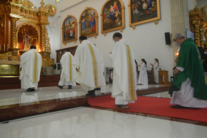 Eine Gruppe von Priestern geht einen roten Teppich in einer Kirche entlang, mit Menschen, die auf dem Boden vor und rechts von ihnen stehen, und Foto Rahmen an der Wand im Hintergrund.