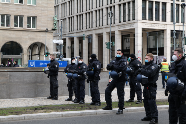 Polizeibeamte in schwarzen Uniformen und Masken stehen vor einem Gebäude mit Glasfenstern und Säulen in Berlin, Deutschland, einige halten Helme, mit Laternenmasten, Verkehrsampeln, Schildern mit Text, einer Statue und anderen Menschen in der Szene.