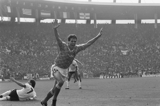 Ein Schwarz-Weiß-Foto eines Fußballspielers, der nach einem Torjubel von einer Gruppe von Menschen auf dem Boden in einem Stadion mit Brettern, Fahnen, Pfosten und einem klaren Himmel im Hintergrund umgeben ist.