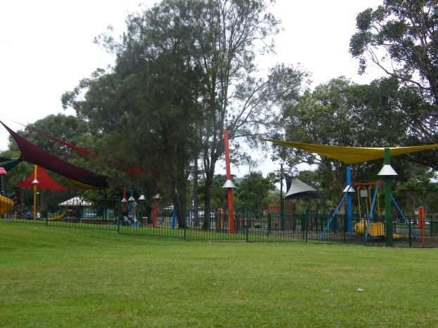 Ein Park mit grünem Gras, umgeben von einem Zaun, mit bunten Spielplatzgeräten wie Schaukeln und Rutschen, vor tallen Bäumen und einem hellblauen Himmel.