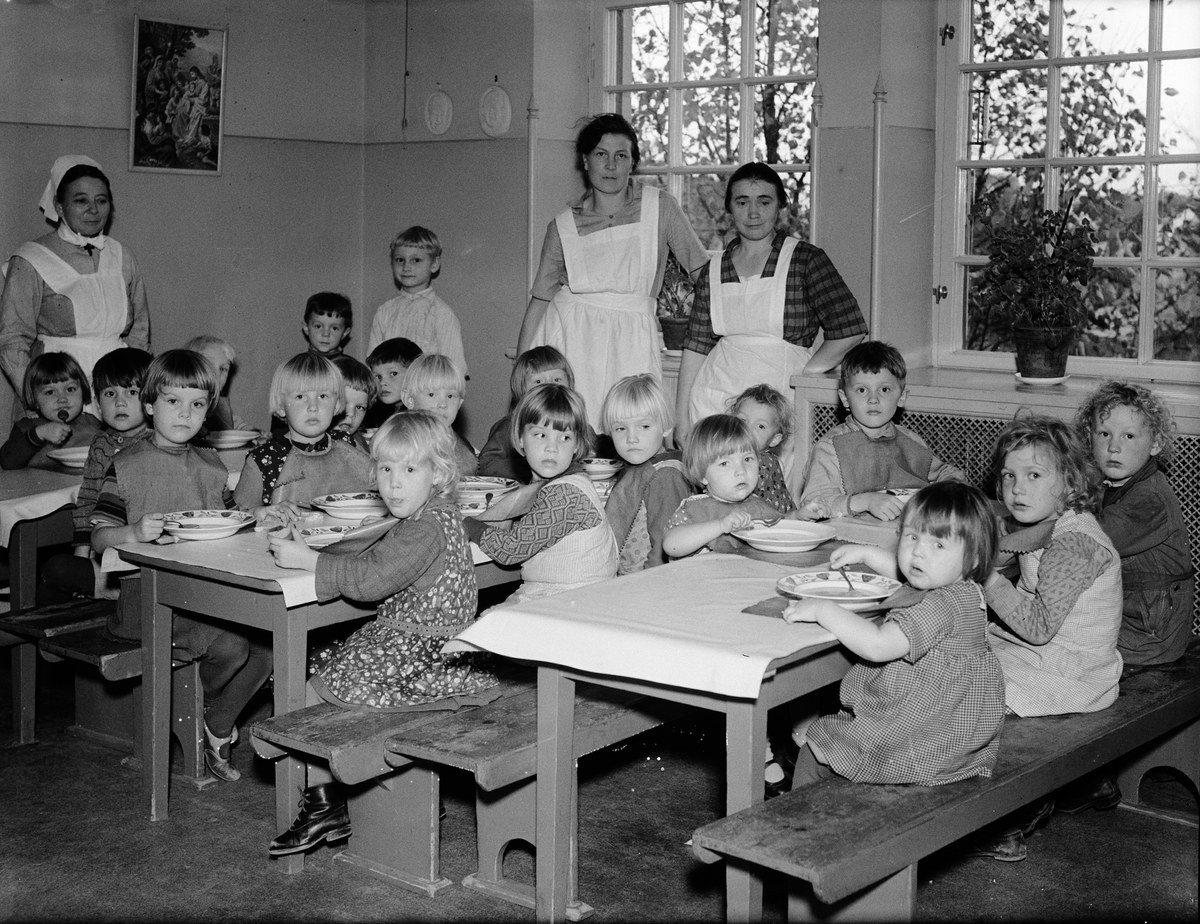 Eine Gruppe von Kindern sitzt an Tischen in einem Klassenzimmer und isst von Tellern, mit Bänken um die Tische und einem gerahmten Foto an der Wand dahinter; Fenster mit Pflanzen sind rechts zu sehen.