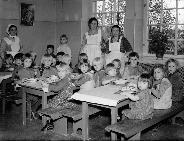 Eine Gruppe von Kindern sitzt an Tischen in einem Klassenzimmer und isst von Tellern, mit Bänken um die Tische und einem gerahmten Foto an der Wand dahinter; Fenster mit Pflanzen sind rechts zu sehen.