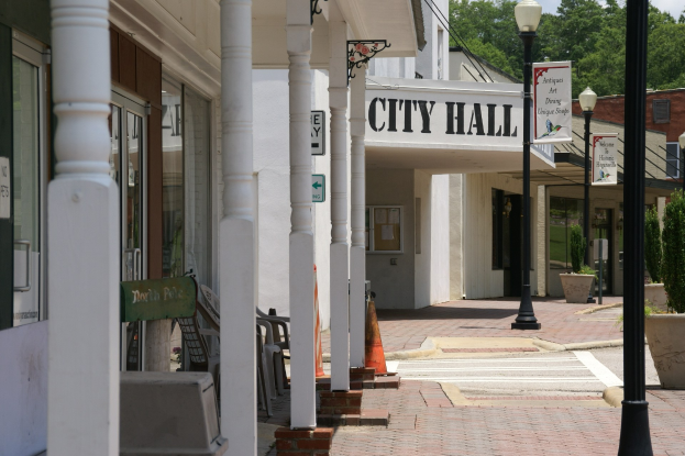 Rathaus mit umliegenden Gebäuden, Straßeninfrastruktur, Fahrzeugen, Verkehrskegeln, Möbeln, Pflanzen und wolkigem Himmel.