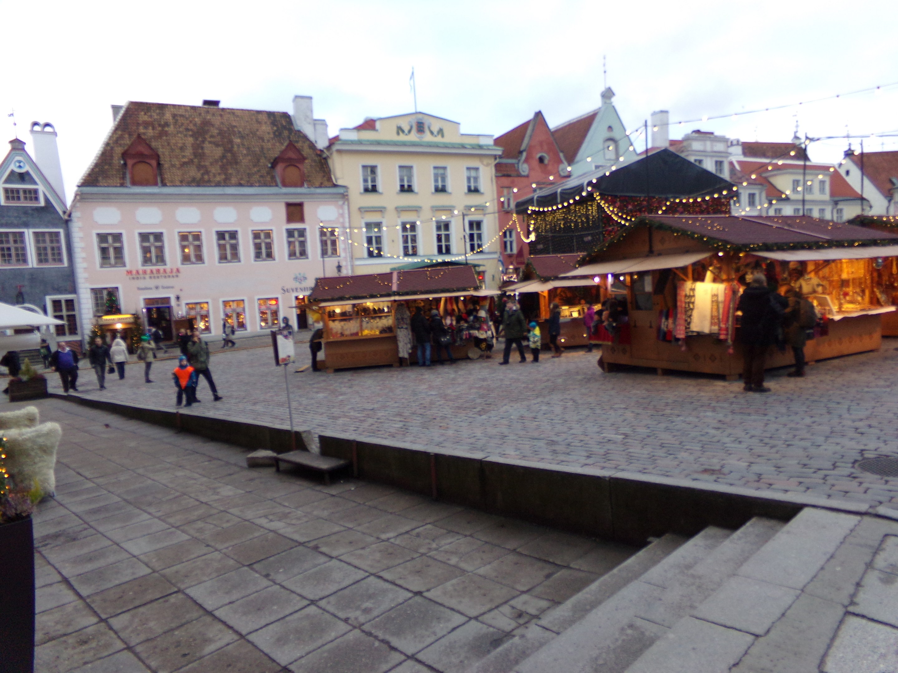 Ein belebter Weihnachtsmarkt in Tallinn, Estland mit Menschen um geschmückte Stände, festliche Gebäude, bewölkter Himmel, Treppe nach oben und Topfpflanzen.