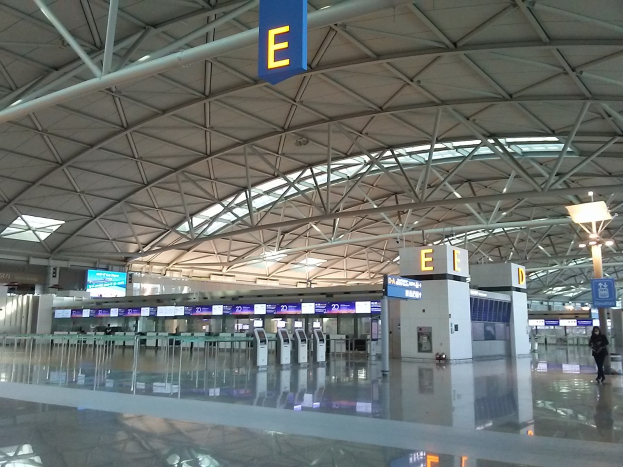 Leerer Flughafen-Terminal mit Menschen, die gehen, Schilder mit Text, Maschinen vorne und Metallstangen und Lampen an der Decke.