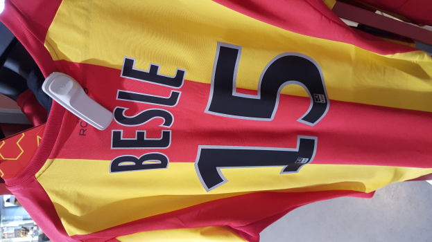 Rotes und gelbes Fußballtrikot mit aufgedrucktem Text und Nummern, das an einer Ladengestell hängend mit anderen Gegenständen und dem Boden im Hintergrund zu sehen ist.