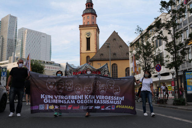Eine Gruppe von Menschen in Masken marschiert eine Straße entlang und hält ein Banner hoch, mit einem geparkten Auto auf der linken Seite und Gebäuden, Bäumen, Schildern, Polen und einem Uhrenturm im Hintergrund unter einem klaren blauen Himmel.