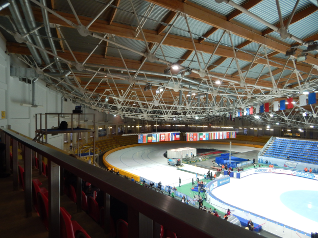 Große Indoor-Eisfläche mit zahlreichen Menschen, die Schlittschuh laufen, umgeben von Sitzplätzen, Geländern, Brettern, Beleuchtungskörpern und strukturellen Elementen, mit einer Wand im Hintergrund.