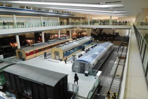 Ein großer Museumsraum mit verschiedenen Zügen in unterschiedlichen Größen und Farben, mit Menschen, die sie vom Boden, Geländern und Säulen unter Deckenlampen betrachten.