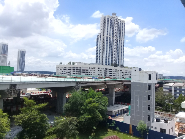Luftaufnahme einer Stadt mit Gebäuden, Bäumen, Gras, Fahrzeugen, einer Brücke, Masten und Wolken, die eine Baustelle für einen neuen Wohnkomplex zeigt.