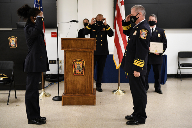 Gruppe uniformierter Personen auf einem Podium mit einem Mikrofon, die eine Urkunde während einer Vereidigung halten, umgeben von Fahnen, Stühlen, Tischen und einer Wand.
