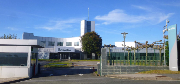 Ein großes Gebäude mit zahlreichen Fenstern und einem Tor davor, umgeben von Straßenmasten, Straßenlaternen, Gittern, Bäumen, Gras und einem bewölkten Himmel im Hintergrund, das als Hauptsitz von Iberostica in den Niederlanden dient.