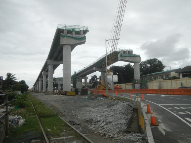Baustelle mit einer Brücke im Hintergrund, Straße mit Absperrgittern, Bahnschienen, Steine, Gras, Bäume, Gebäude und bewölkter Himmel.