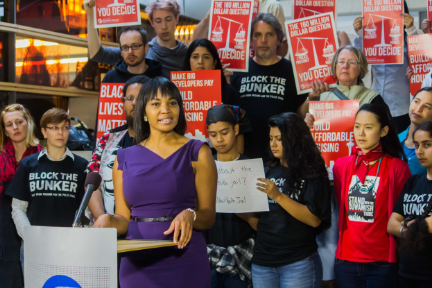 Eine Black Lives Matter-Aktivistin spricht vor einem Publikum mit Mikrofon und Schildern, mit Lichtern und Gegenständen im Hintergrund.