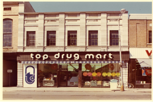 Eckladen namens "Top Drug Mart" mit einem Schilder, Fenstern, einem Straßenpfosten, einem geparkten Fahrrad, einem Müllcontainer und einem bewölkten Himmel.