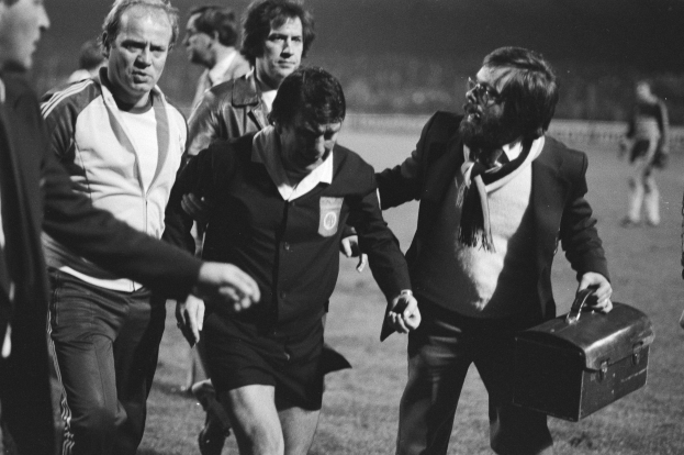 Schwarzes und weißes Bild einer Gruppe von Männern, die auf einem Fußballfeld gehen, einer hält eine Aktentasche, die Männer sehen besorgt aus.