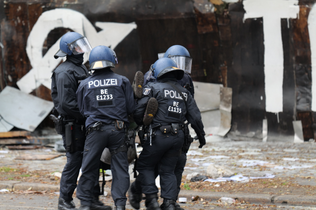 Polizeibeamte in schwarzen Uniformen und blauen Helmen stehen vor einem Gebäude mit Graffiti während einer Ausschreitung, mit verstreuten Papieren und Gegenständen auf dem Boden.