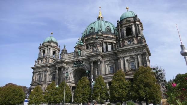 Großes, prunkvolles Berliner Dom-Gebäude in Berlin, Deutschland, mit architektonischen Details wie Statuen und Bögen, vor einem bewölkten Himmel, mit Passanten, Bäumen und einem Turm im Vordergrund.
