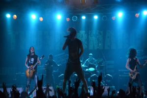 Eine Gruppe von Musikern performs auf einer hell erleuchteten Bühne, wobei einige Gitarren spielen und einer ein Mikrofon hält, während ein Drummer im Hintergrund spielt und die erhobenen Hände des Publikums am Boden sichtbar sind.