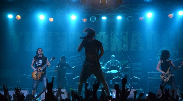 Eine Gruppe von Musikern performs auf einer hell erleuchteten Bühne, wobei einige Gitarren spielen und einer ein Mikrofon hält, während ein Drummer im Hintergrund spielt und die erhobenen Hände des Publikums am Boden sichtbar sind.