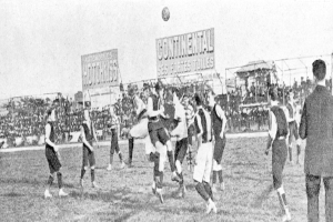 Ein Schwarz-Weiß-Bild einer Gruppe von Männern, die auf einem Feld Füßball spielen, mit einem Ball in der Luft und Zuschauern im Hintergrund, mit Tafeln, die Text über das Spiel anzeigen.