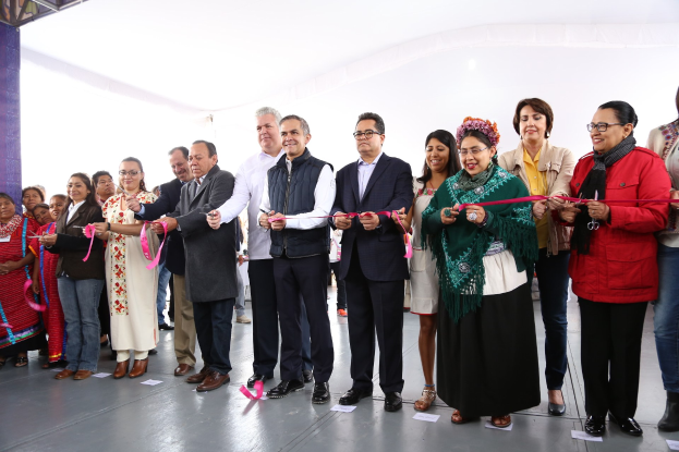 Eine Gruppe von Menschen bei einer Scherenschnitte-Zeremonie, mit einer Säule links daneben, verstreuten Papieren auf dem Boden und einem weißen Tuch im Hintergrund.