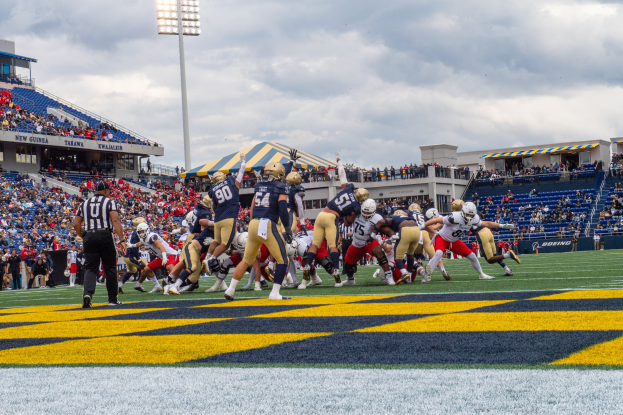 Ein Fussballspiel im Gange mit Spielern in Sportbekleidung und Helmen im Vordergrund, Zuschauern, Geländern, Beschriftungen, Laternen und einem bewölkten Himmel im Hintergrund sowie Text am unteren Bildrand.