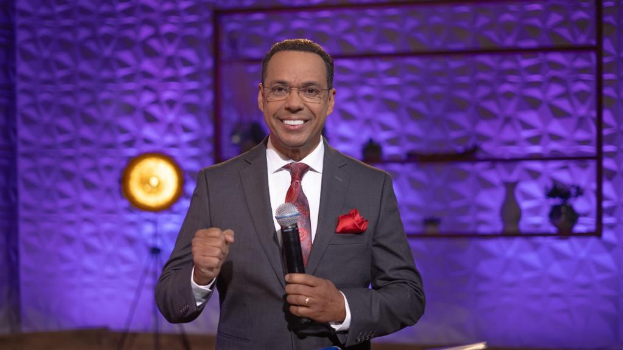 Stephen Colbert, Moderator von "The Daily Show with Trevor Noah", steht auf einer Bühne und hält ein Mikrofon in der Hand, trägt einen Anzug, eine Krawatte und eine Brille, hinter ihm ist ein Licht und ein Ständer zu sehen.