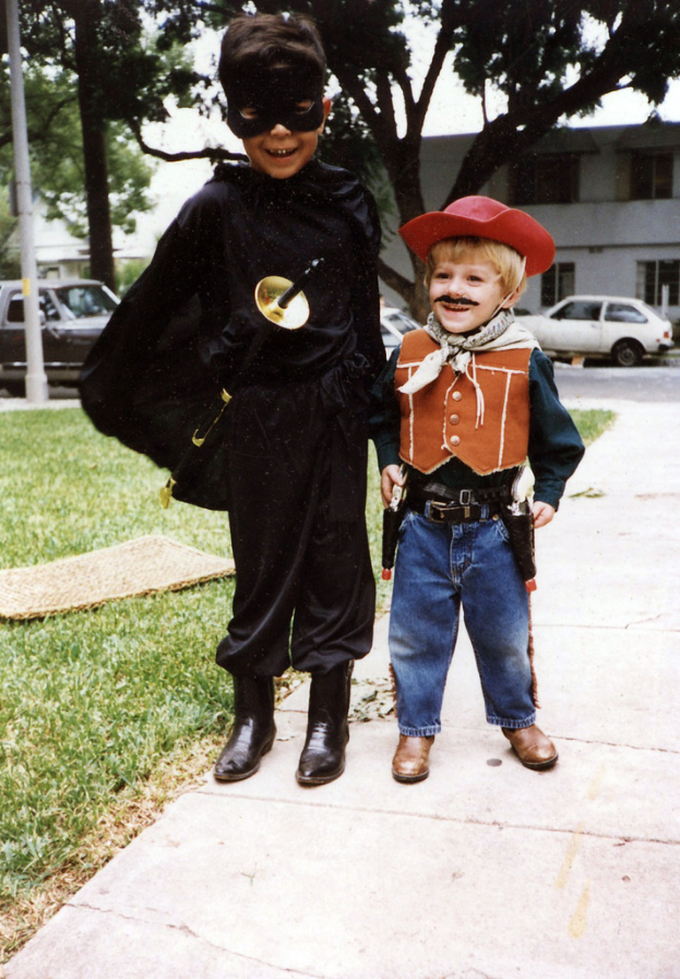 Kinder in verschiedenen Kostümen auf einer Straße mit Fahrzeugen, Gebäuden, Bäumen und Straßenlaternen im Hintergrund unter einem Himmel.