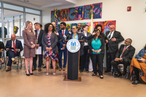 Eine Gruppe von Menschen, die vor einem Podium mit Mikrofon und einer Tafel mit der Aufschrift "Sancho City Councilman Joins City Council" stehen und sitzen, mit Flaggen, Feuerlöscher und Deckenleuchten an der Rückwand.